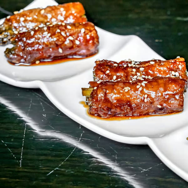 Two white plates of Agami Sushi's teriyaki-glazed beef rolls with green onions and sesame seeds.
